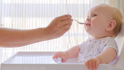 Wall Mural - Mother is Feeding a baby from a spoon. Mom feeds a 9 month baby with fruit puree from a spoon, close-up, high key. Infant boy eats sitting on baby's chair. Mother cares about little son.