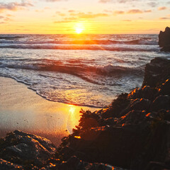 Poster - Beautiful natural landscape with sandy beach  with big stones during sunset.  Sun over horizon and Rolling ocean waves to the rocky shore while sunset.  Summer tourist destination in Portugal. Travel