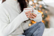 A beautiful brunette girl in a white sweater and blue jeans sits on a bed with a cup of hot drink in her hands
