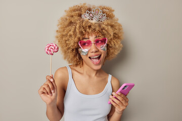 Wall Mural - Funny woman with curly bushy hair holds caramel candy on stick and modern smartphone exclaims from joy applies silver patches under eyes wear small crown t shirt isolated over grey background