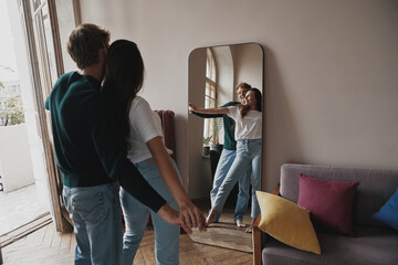 Wall Mural - Playful young couple holding hands while looking at their reflection in the mirror at home