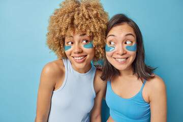 Canvas Print - Horizontal shot of mixed race women apply beauty patches under eyes to reduce puffiness undergo skin care treatment dressed in casual t shirts have good mood isolated over blue background. Wellness