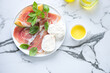 Plate with prosciutto ham, cantaloupe, mozzarella and green basil, top view on a white marble background, horizontal shot