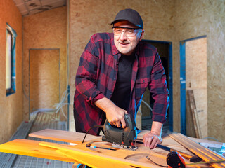 Man is carpenter. Guy with electric jigsaw is engaged in woodworking. Carpenter smiles and looks into camera. Man carves wooden elements. Carpenter at work. Worker inside house under construction