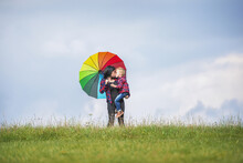 A Little Boy In A Checkered Shirt And Blue Pants With His Mom Kiss On Top Of A Meadow Near The Horizon. Behind Them The Sky Is Dotted With Clouds. A Woman Is Holding A Colorful Umbrella In Her Hand.
