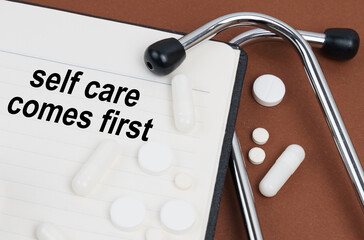 On a brown surface lies a stethoscope, pills and a notepad with the inscription - self care comes first