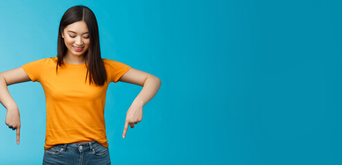 Cute tender feminine asian girl dark haircut look pointing down contemplating interesting product, stare curiously smiling amused, express enthusiasm interest try-out cool thing, blue background