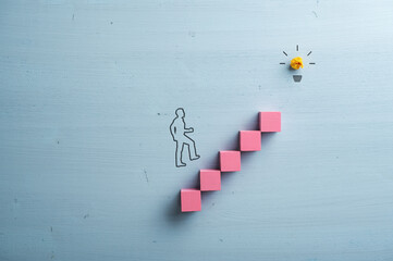 Wall Mural - Silhouette of a businessman walking up the stairs made of pink wooden blocks leading towards a glowing light bulb