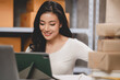 © chokniti - online shopping concept, happy young Asian business owner woman prepare parcel box and check orders of product for deliver to customer on laptop computer at home, e-business marketing on cyberspace