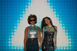 © Clique Images - Studio portrait of two ethnically diverse young women wearing disco fashion outfits standing in projector light holding hands looking at camera