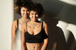 © Clique Images - Horizontal medium multiple exposure studio shot of attractive young Black woman with tattoos on body wearing lounge bra standing in sunlight smiling at camera