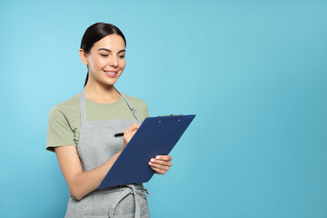 Poster - Young woman in grey apron with clipboard on light blue background, space for text