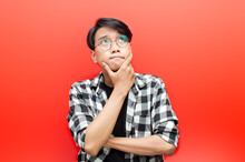 Thoughtful Asian Man Thinking And Wondering With Hand Touching Skin As Thinking Gesture Isolated Over Red Background.