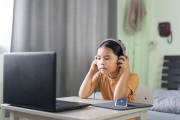 Wall Mural - Asian child or kid girl e-learning listening by studying online class on computer notebook with wearing headphone or headset for video call communication or person learn from home school by laptop