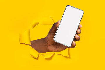 African american male hand holding smartphone with empty screen showing device breaking through yellow paper sheet