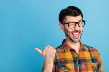 Wall Mural - Photo of friendly handsome positive guy in eyewear plaid shirt indicating at empty space blinking isolated on blue color background