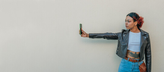 black latin girl looking at mobile phone with outstretched arm and background wall in the street outdoors