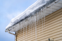 Sharp Spiked Icicles On Roof With Snow Against Blue Sky. Icicles On Roof Of Private House Resulting From Improper Construction Of Roof. Metal Downpipe System, Guttering System At Winter