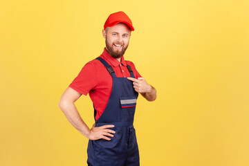 Wall Mural - Side view of proud successful worker man standing and pointing at himself, bragging about the result of his work, wearing overalls and red cap. Indoor studio shot isolated on yellow background.