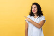 Portrait of woman with dark wavy hair pointing finger away with toothy smile, wondering about empty space for your advertisement, template. Indoor studio shot isolated on yellow background.