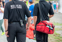 Medical First Responders Walking Along A Road Wearing Black Uniforms, With Medical First Responder In Grey Letters Across The Back Of The Paramedic. The EMT Is Carrying A Red First Aid Kit And Radio.