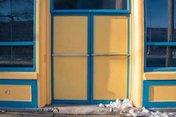 Two bright yellow doors with chrome handle to a store with blue trim. Over the door is a transom glass window with multiple panes. There's a sidewalk in front of the tall entrance to the vintage shop 