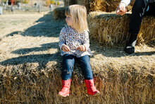 Little Girl In Cowgirl Boots Sitting On A Bale Of Hay