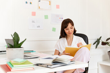 Beautiful young caucasian woman makes entries in diary sitting at her desk. Brunette wears glasses, white blouse. Focused on work concept