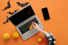 From Above Flat Lay Shot Of Unrecognizable Woman Sitting At Orange Table With Pumpkins, Paper Bats On It In Front Of Laptop Buying Things For Halloween Online