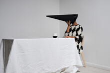 Horizontal Studio Shot Of Mysterious Young African American Woman Wearing Black And White Dress With Creepy Conus-shaped Mask On Face Sitting At Table