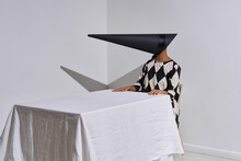 Horizontal Studio Shot Of Modern Young African American Fashion Model Wearing Black And White Outfit With Creepy Conus-shaped Mask On Face Sitting At Table
