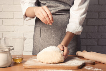 Woman sprinkling flour over dough at table near white brick wall, closeup