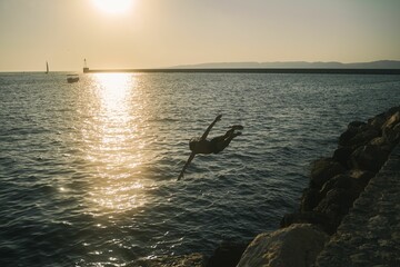 Poster - A person jumping down, high diving from the cliff, rocks, to the ocean during sunset