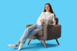 © Pixel-Shot - Beautiful woman reading book in armchair on blue background