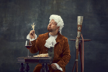 Strange things. Vintage portrait of young man in brown vintage suit and white wig like medieval royal hunter isolated on dark background.