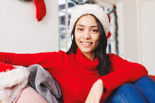 Portrait Of Happy Biracial Woman Wearing Santa Hat, Sitting On Sofa In Living Room
