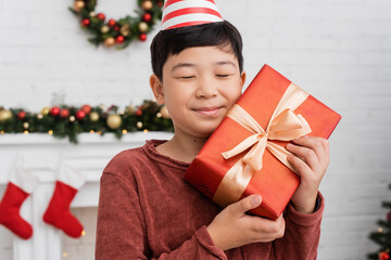 Wall Mural - Happy asian boy in party cap holding gift near blurred christmas decor at home