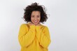 © Roquillo - Satisfied young beautiful woman with curly short hair wearing yellow sweater over white background touches chin with both hands, smiles pleasantly, rejoices good day with lover