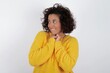 © Roquillo - Happy young beautiful woman with curly short hair wearing yellow sweater over white wall anticipates something awesome happen, looks happily aside, keeps hands together near face, has glad expression.