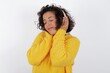 © Roquillo - Pleased young beautiful woman with curly short hair wearing yellow sweater over white backg enjoys listening pleasant melody keeps hands on stereo headphones closes eyes. Spending free time with music