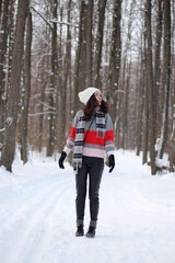 Wall Mural - A young woman in a sweater and a white hat in the forest. Full length portrait. Snowing. Winter atmosphere. Walk in the forest.