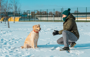 Wall Mural - Young man blowing snowflakes from his hands to a dog golden retriever in a winter day. Friendship, pet and human.