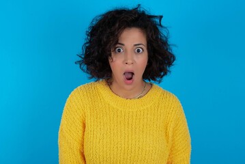 Wall Mural - Expressive facial expressions. Shocked stupefied young beautiful woman with curly short hair wearing yellow sweater over blue wall, keeps jaw dropped feels stunned from what he sees aside.