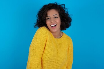 Sticker - young beautiful woman with curly short hair wearing yellow sweater over blue wall with broad smile, shows white teeth, feeling confident rejoices having day off.