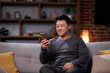 © YURII MASLAK - Smiling Asian man communicates in social networks and uses a smartphone. A man in casual clothes looks through a mobile phone, communicates in a friendly way, sits on a sofa and relaxes at home.