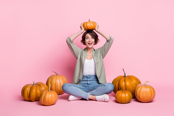 Wall Mural - Full body photo of young cheerful girl have fun look pumpkin on head organic sit floor isolated over pink color background