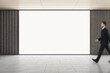 Leinwandbild Motiv Advertising concept with businessman walking by blank white glowing screen with space for your logo or text in empty hall with light wooden ceiling and grey concrete floor, mockup