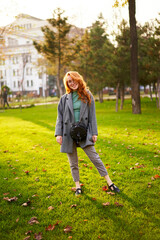 Redhead smiling pretty woman walking in park on sunny autumn day. Elegant female poses in stylish authentic outfit has fun laughing on green lawn with vivid foliage, leaves. Girl enjoys fall season.