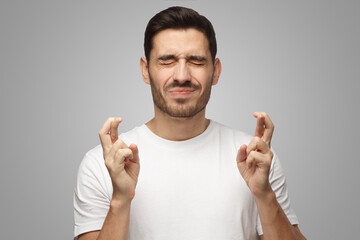 Wall Mural - Closeup of man with closed eyes in white t-shirt on gray background crossing fingers for luck