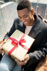 happy african american man in coat sitting with gift box outdoors.